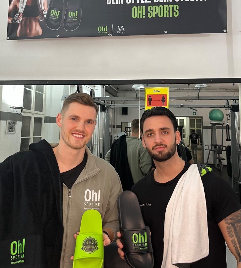 Übergabe Oh Sports Zwei Männer in Trainingskleidung stehen lächelnd vor einem Fitnessstudio-Schild.